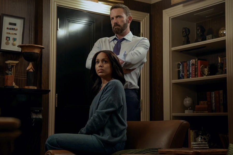 "A woman sits on a leather chair looking concerned, while a man stands behind her with arms crossed in a dimly lit office or library filled with shelves, books, and decorative objects."