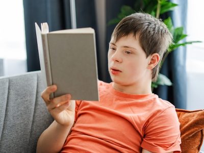 Young man with down syndrome enjoying a book, comfortably sitting on a sofa at home, student studying. Knowledge concept, education